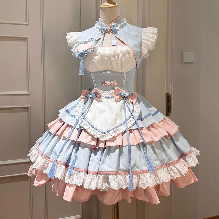 a full view of a frilly blue and pink lolita fashion dress is shown on a mannequin against a white and gold wall.  the bodice of the dress is white with pink accents and blue bows. the skirt is full and tiered, with layers of blue and pink fabric. the dress is decorated with lace and ribbons.