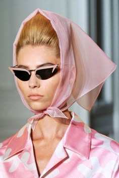 a beautiful woman with blonde hair is wearing pink sunglasses and a pink scarf that wraps around her head. she is looking directly at the camera. she has a casual and vintage style that is reminiscent of the 1950s and 1960s. she is wearing a pink blouse with a white floral pattern and a pink scarf that is tied around her head. the background is blurred.