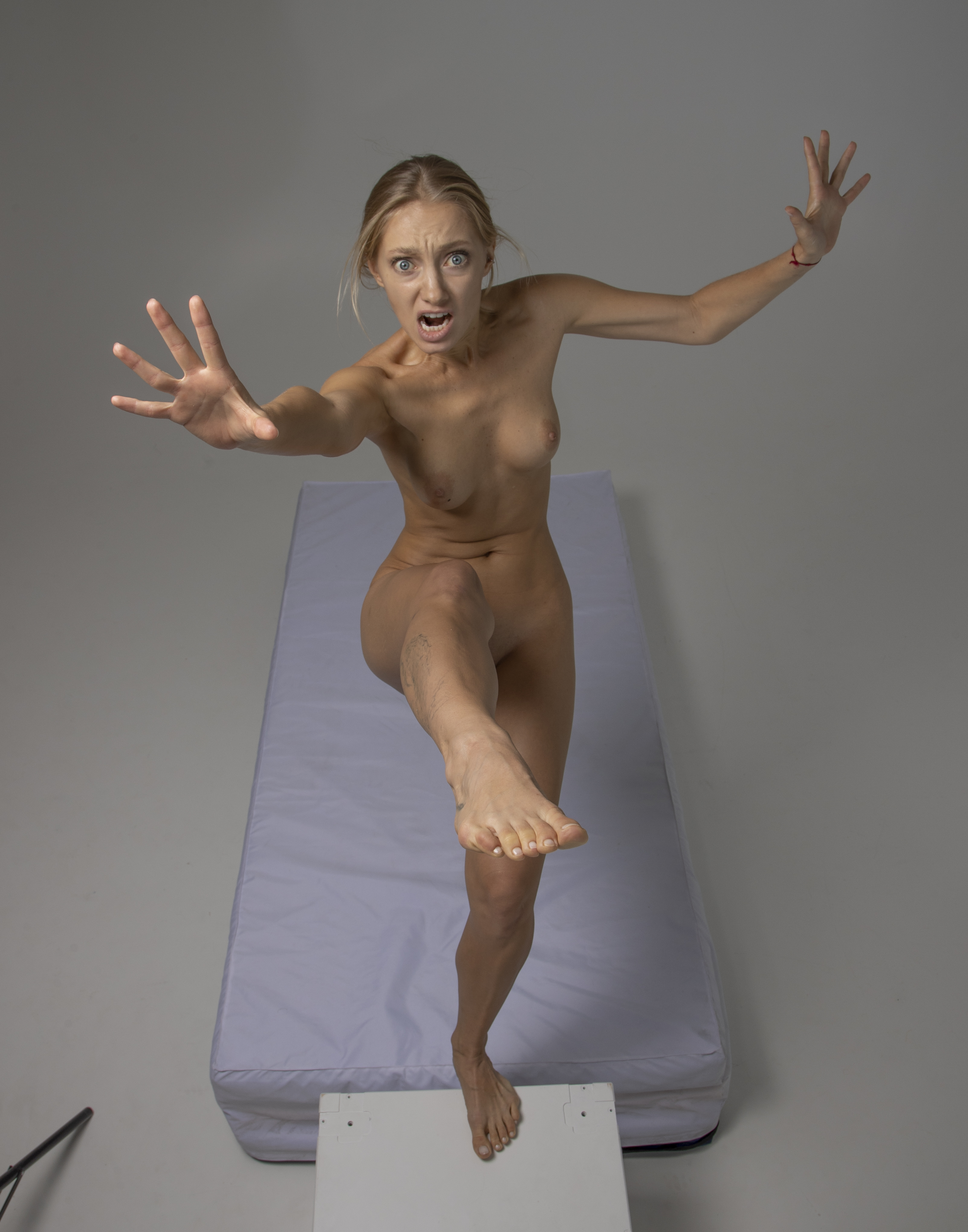 a fullbody studio shot of a nude woman expressing fear and panic. she stands on a small white platform positioned next to a mattress, her arms outstretched and hands open as if warding something off. her face is contorted in a scream, eyes wide with terror. she has bare feet, and the gray background emphasizes her figure.