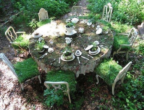 an image of an old, circular table covered in moss and vines. the table is set with teacups, silverware and plants growing out of teapots.  surrounding the table are chairs, also covered in moss and vines, giving the impression of being abandoned long ago.