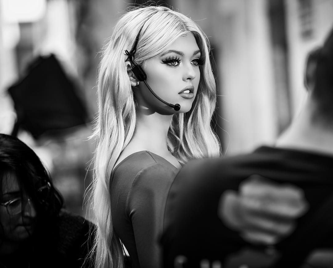 a black and white portrait of a blonde woman wearing a headset with a microphone. she is looking off to the side with a slightly open mouth. the background is blurred, and there are other people partially visible in the foreground and background.