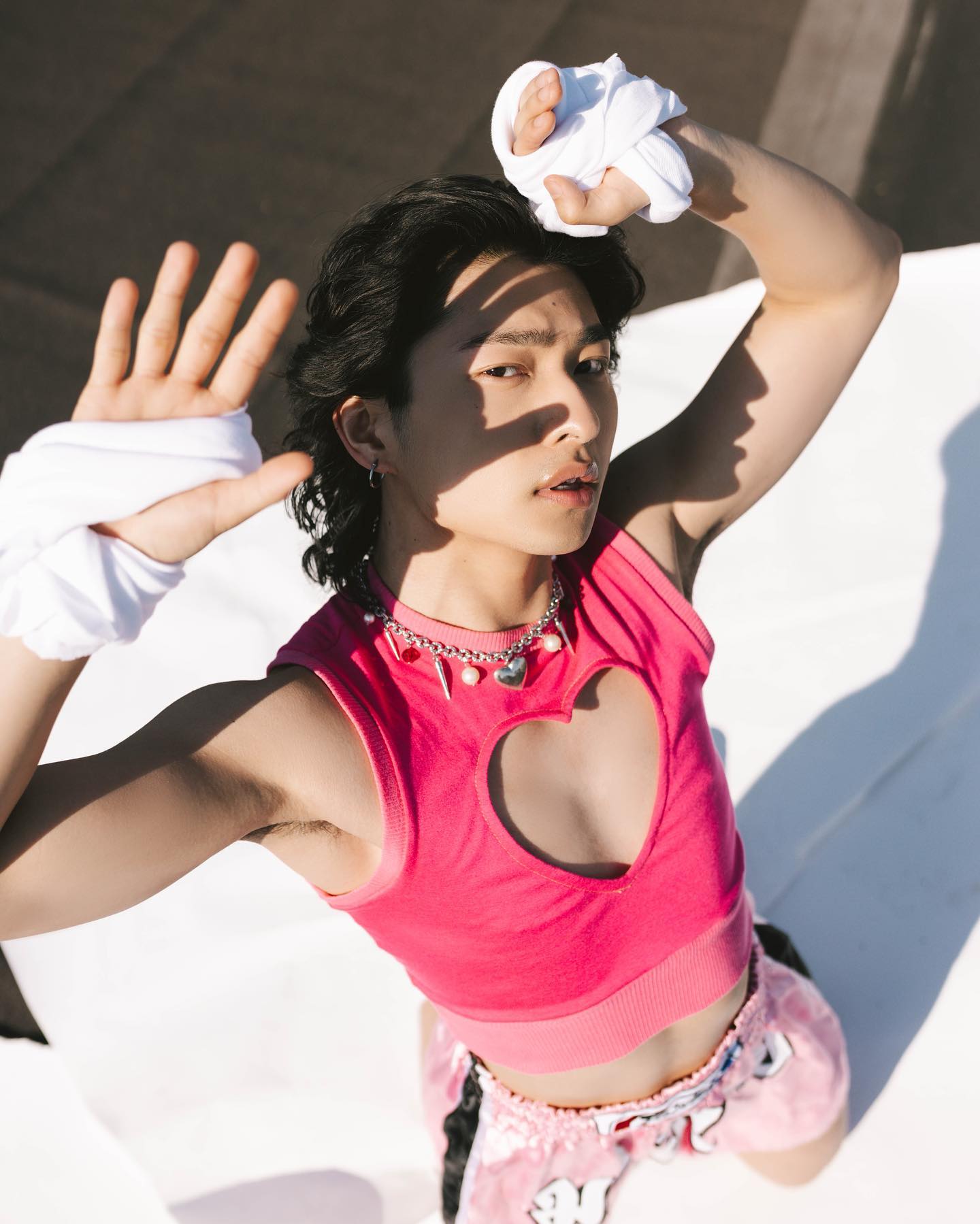 a young asian person with short dark hair is posing on a white background outdoors in the bright sunlight.  they are wearing white hand wraps, a pink heartcutout crop top, and pink and white boxing shorts. the high camera angle looks down at the subject who is looking directly at the camera and shielding their eyes with one hand. the bright sun casts harsh shadows across their face and body. the overall aesthetic is playful and blends traditionally masculine and feminine elements.
