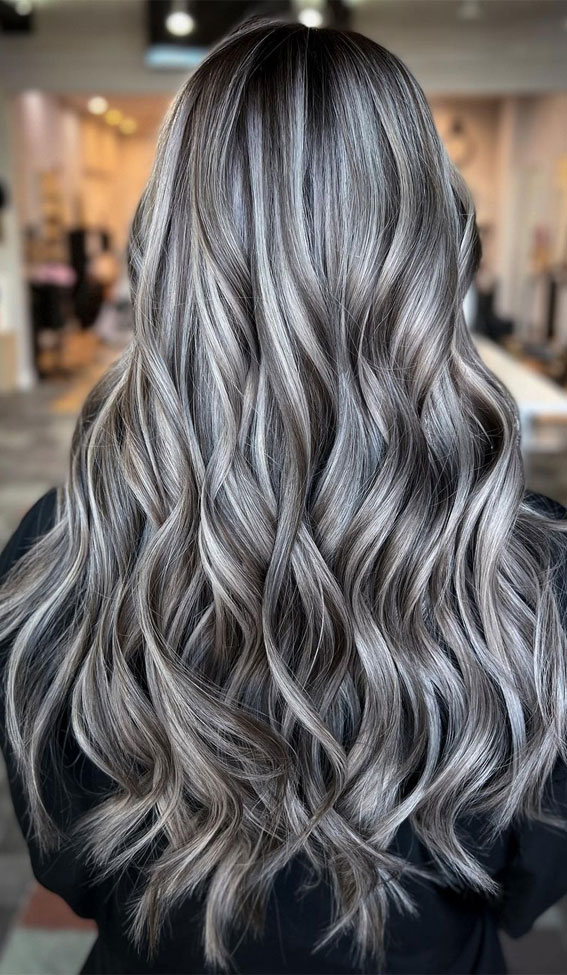 he image showcases a woman with long, wavy silver hair with darker gray highlights. she is standing indoors, likely in a hair salon, as suggested by the blurred background. the lighting is bright, illuminating her hair and emphasizing the shine and texture. the image is taken from a rear view, focusing solely on the woman's hair and not showing her face.