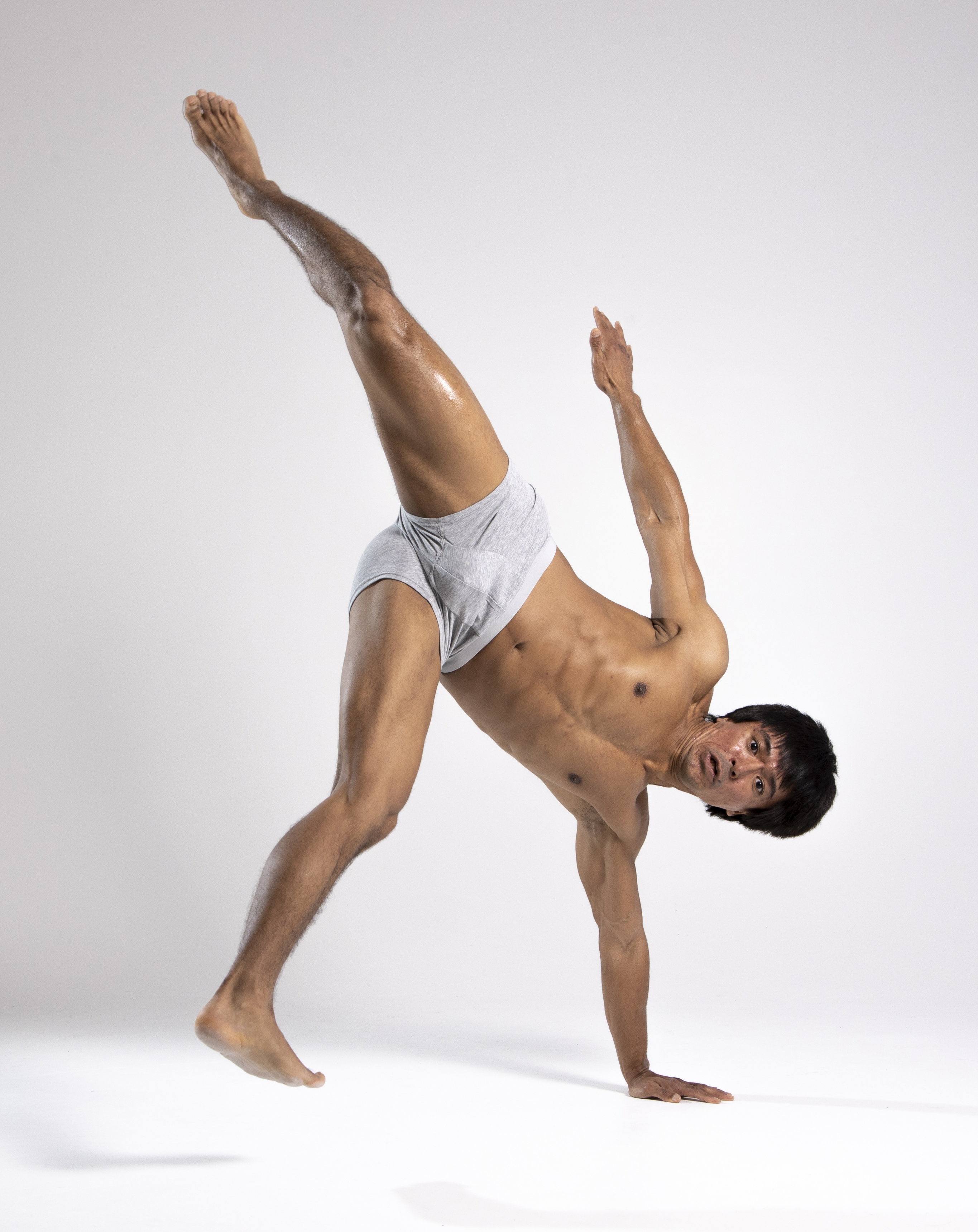 a muscular man wearing only gray underwear is captured midmovement, performing a onehanded handstand in a studio setting. one leg is extended high into the air, while the other leg is bent and provides partial support. the man is shirtless, highlighting his physique, and his face shows a concentrated expression. the white background emphasizes the athleticism and balance of the pose.