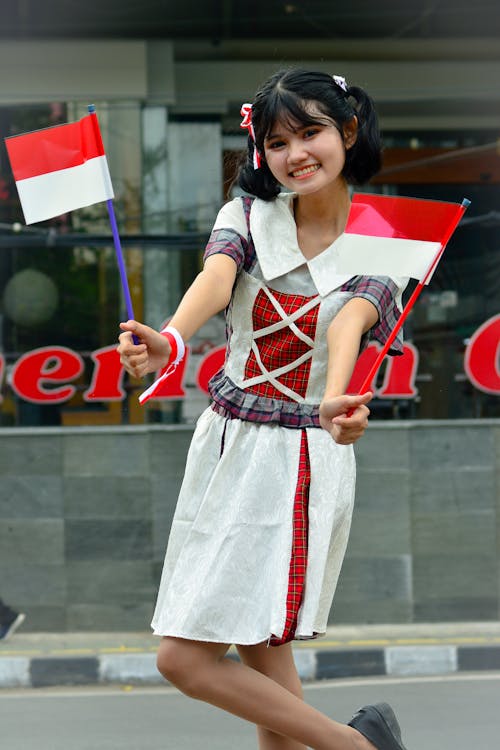 a woman in a dress is smiling and holding two flags of indonesia in a medium shot outdoors. the woman has pigtails and bangs.