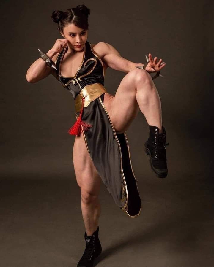 a muscular woman in a chunli cosplay outfit poses in a studio. she is wearing a black dress with gold trim and red tassels, spiked bracelets, and black boots. she has her hair styled in buns. she is striking a fighting pose with one leg raised and arms in a ready position. the background is a neutral gray.