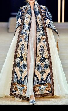 a model walks the runway wearing an elaborately embroidered gown that has black and gold details and a white sheer skirt.  the dress is a beautiful piece of highfashion craftsmanship.