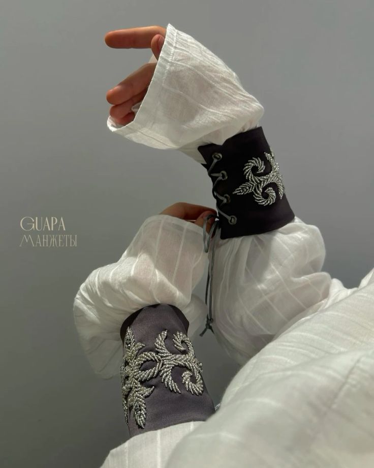 the image showcases a closeup view of a woman's hands wearing a pair of intricately designed cuffs.  the cuffs are dark gray, possibly made of fabric or leather, and feature a striking silvertoned embroidery. the embroidery appears to be beaded and forms a detailed, stylized floral or leaf pattern. the cuffs have a laceup closure at the back. the woman is wearing a loosefitting, longsleeved white blouse, which contrasts beautifully with the dark gray cuffs. the overall style suggests an elegant, possibly vintage or bohemian aesthetic. the brand name "guara" and the word "манжеты" (meaning "cuffs" in russian) are subtly displayed on the image.  the focus is on the detail and craftsmanship of the cuffs and their stylish presentation.