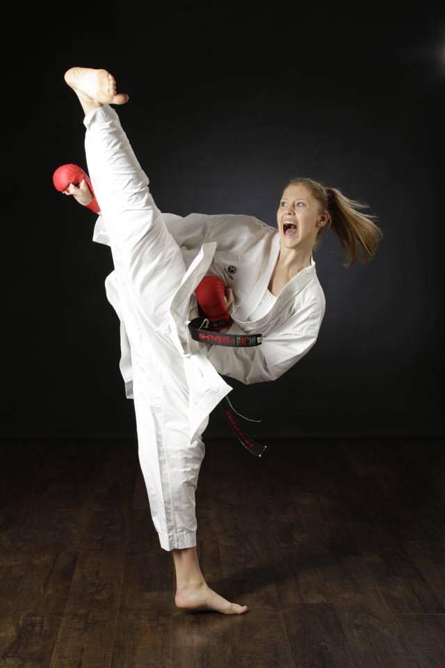 a woman wearing a white karate uniform and a black belt is executing a powerful kick in a studio setting. her leg is extended high in the air, and she is holding a red glove in her hand. her expression is one of focus and determination. the background is dark, and the floor is made of wood.  the photograph captures the intensity and power of martial arts.