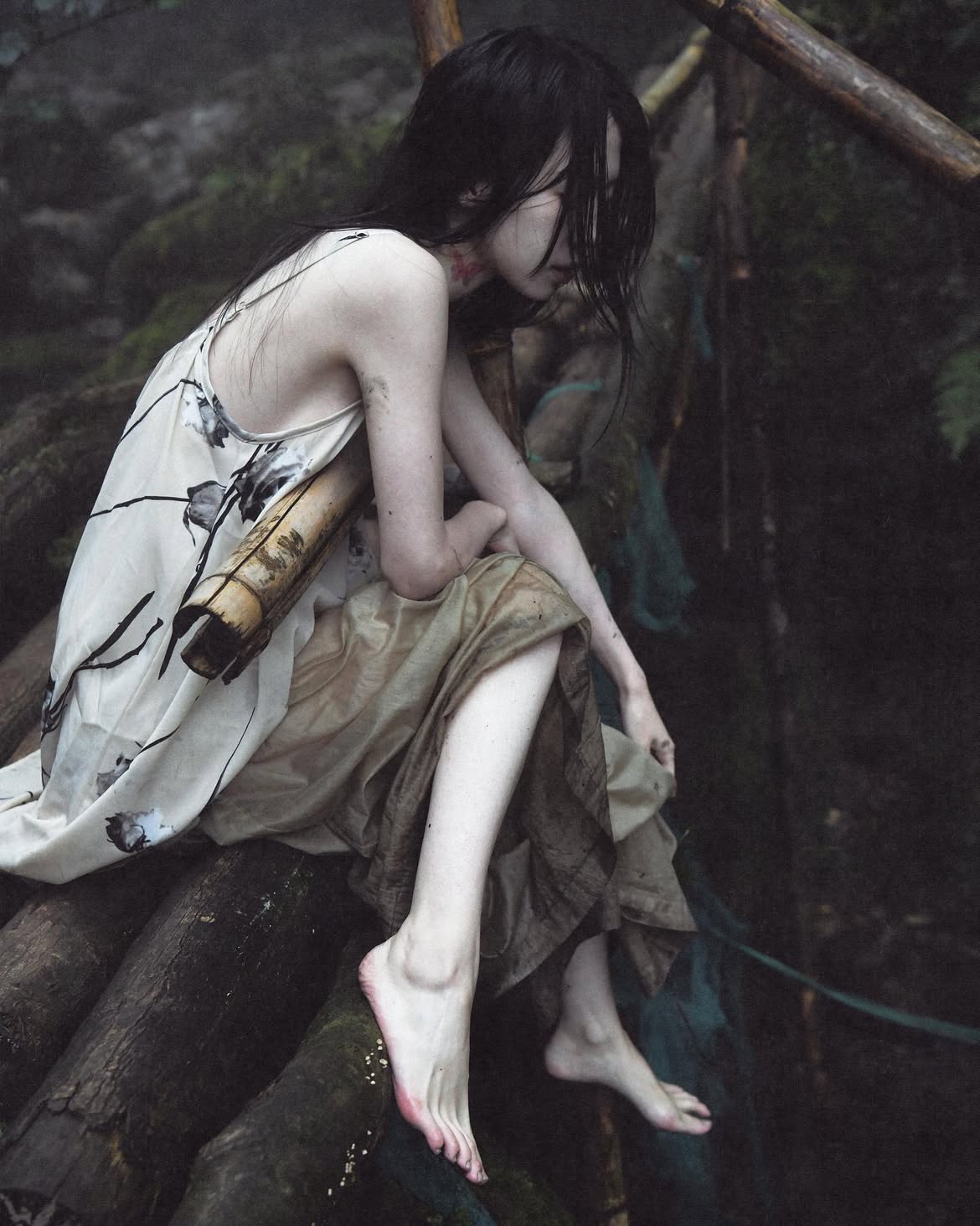 a woman with dark, wet hair is sitting barefoot on some tree trunks in a dark, damp wooded area. she appears pensive and melancholy. she is wearing a lightcolored dress with a pattern and a darker skirt. the scene has a moody, somber atmosphere.