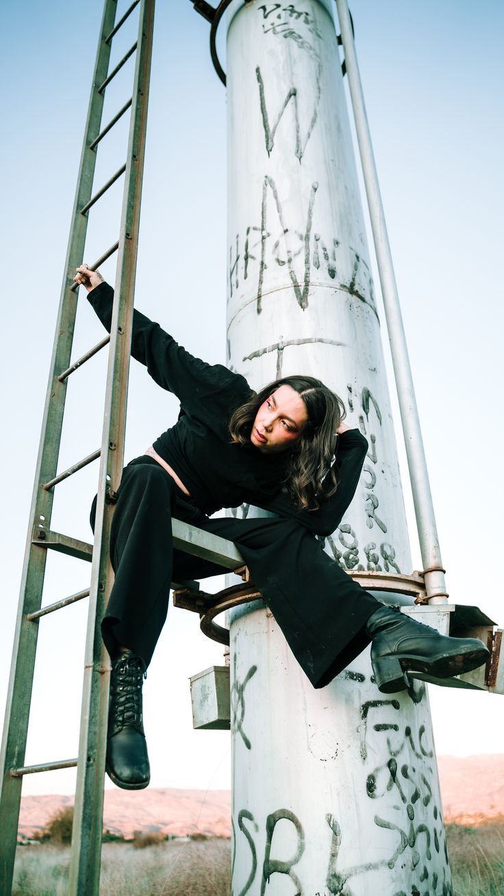 a young woman dressed in a black outfit and boots poses on a ladder and a large metal pole.  the pole has many graffiti markings on it.  the woman is posing with her leg on the pole.  the photo is taken outdoors in a grunge industrial setting.