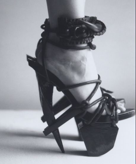 a black and white image shows a foot in an avantgarde high heel. the heel has a pointed shape and the shoe features multiple ankle straps wrapped around the ankle. the overall style is fashionable and striking.