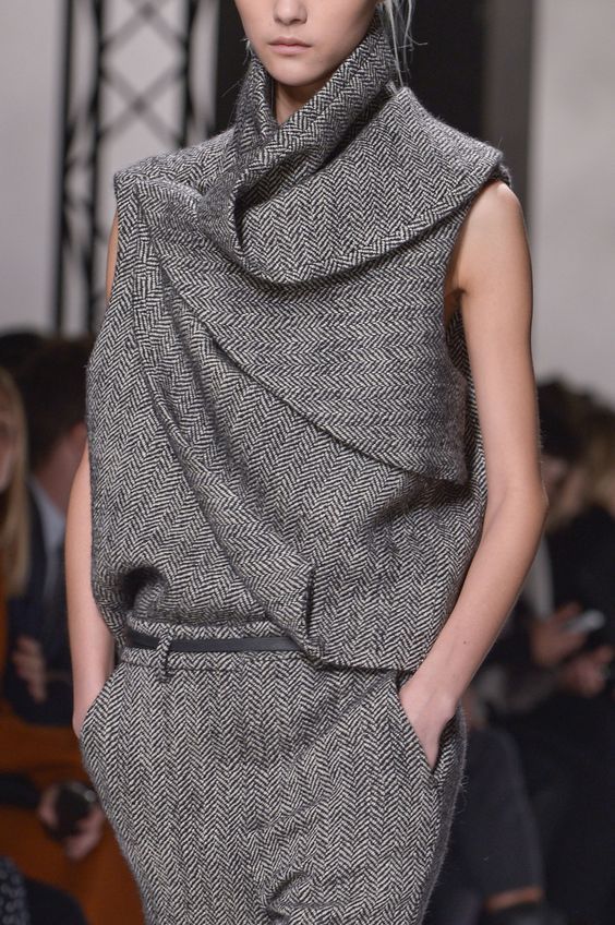 a fashion model is wearing a gray and white herringbone outfit on a runway. the outfit includes a sleeveless top with a high neck and a collar that drapes over itself, and pants that are also herringbone. the model is looking away from the camera and has a serious expression on her face.  the model's arms are at her side, one hand is in her pocket, and the other is hanging down. the outfit is stylish, elegant, and sophisticated.