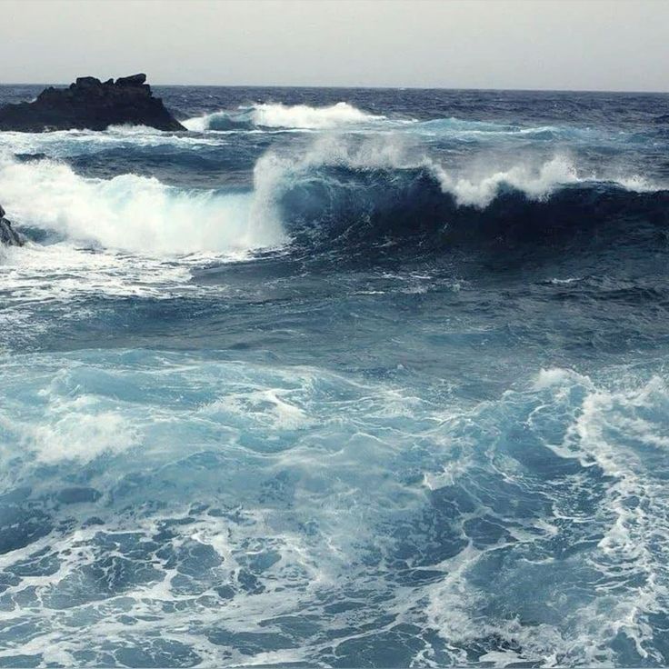 a wide shot of the ocean with large waves crashing against a rocky shore. the water is a deep blue, and the foam from the waves is white. the sky is overcast, and there is a feeling of wildness and power to the image.