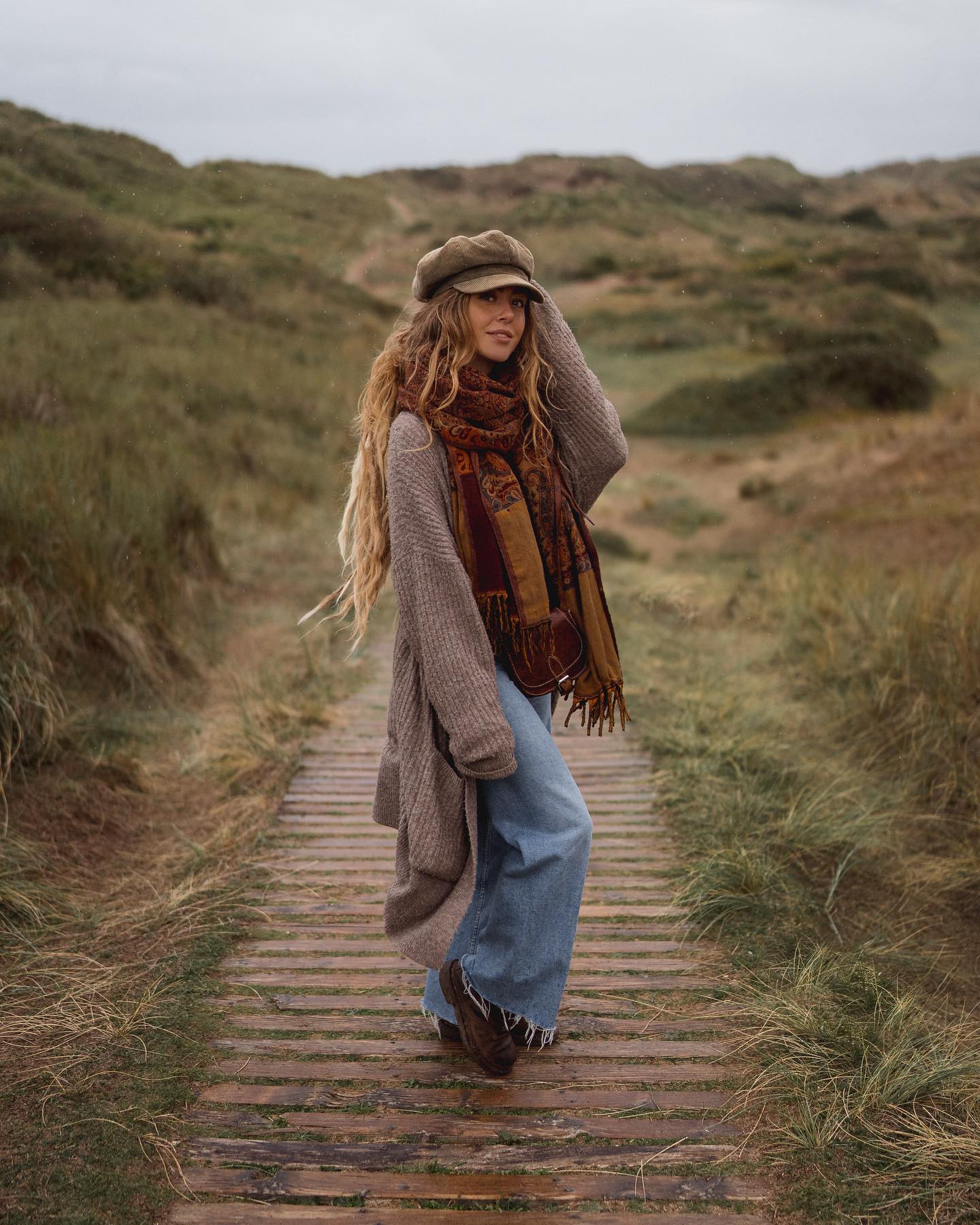 a young woman with long, reddishblonde dreadlocks is standing on a wooden boardwalk that winds through grassy dunes. she's wearing a taupecolored cardigan, wideleg blue jeans, brown boots, a patterned scarf, and a newsboy cap. her style is bohochic, and the overall atmosphere of the image is relaxed and autumnal.  the sky is overcast and the day appears cool.