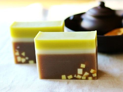 two bars of handmade soap, layered in brown, white and green, with small yellow flecks throughout. the soap bars are sitting on a table next to a rustic ceramic teapot.  the setting is calming and suggests spa treatments or relaxation.