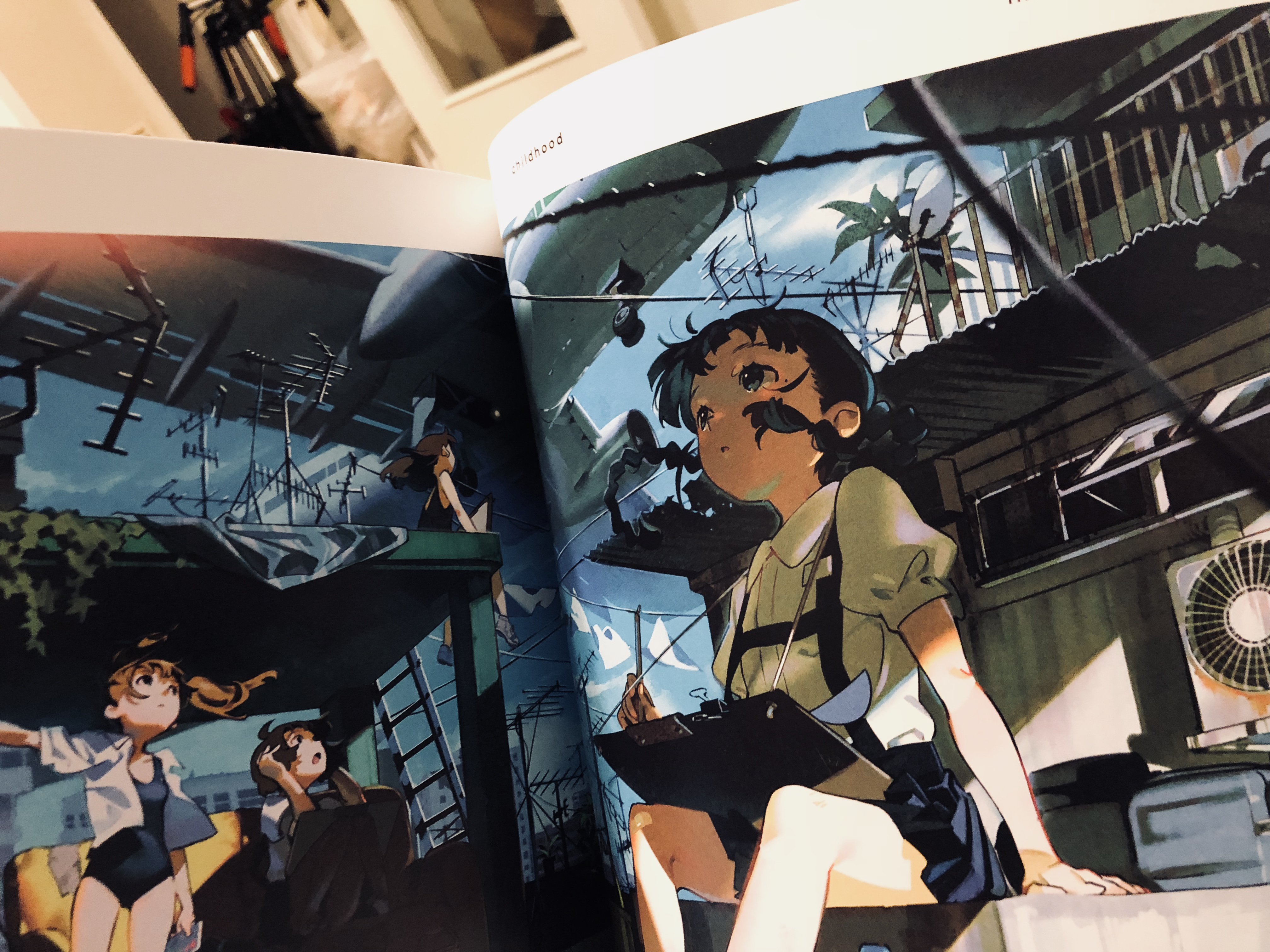 the image shows an open art book featuring an anime illustration. the scene depicts three girls on a rooftop with a low angle perspective, giving the impression of looking up at them. the environment is urban, with buildings and power lines visible in the background. the lighting suggests a bright, sunny day, casting shadows across the scene. the overall color palette is vibrant, with blues and greens dominating. one girl in a white shirt sits prominently in the foreground, while two others stand in the background engaging in conversation.  the illustration has a distinct anime style and appears to be concept art or an illustration from an art book.