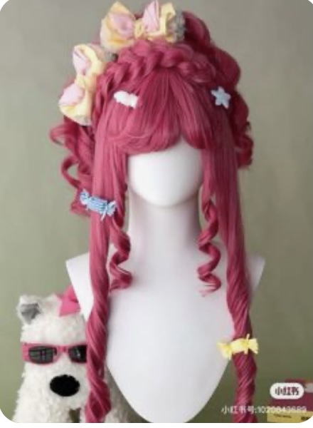 a pink cosplay wig with long, curly locks is displayed on a white mannequin head. the wig features a braided section on top, adorned with yellow and blue bows.  additional small bows and a candyshaped hair tie add to the playful and elaborate style. the mannequin is positioned against a plain backdrop, with a white dog figurine wearing sunglasses partially visible in the lower left corner.