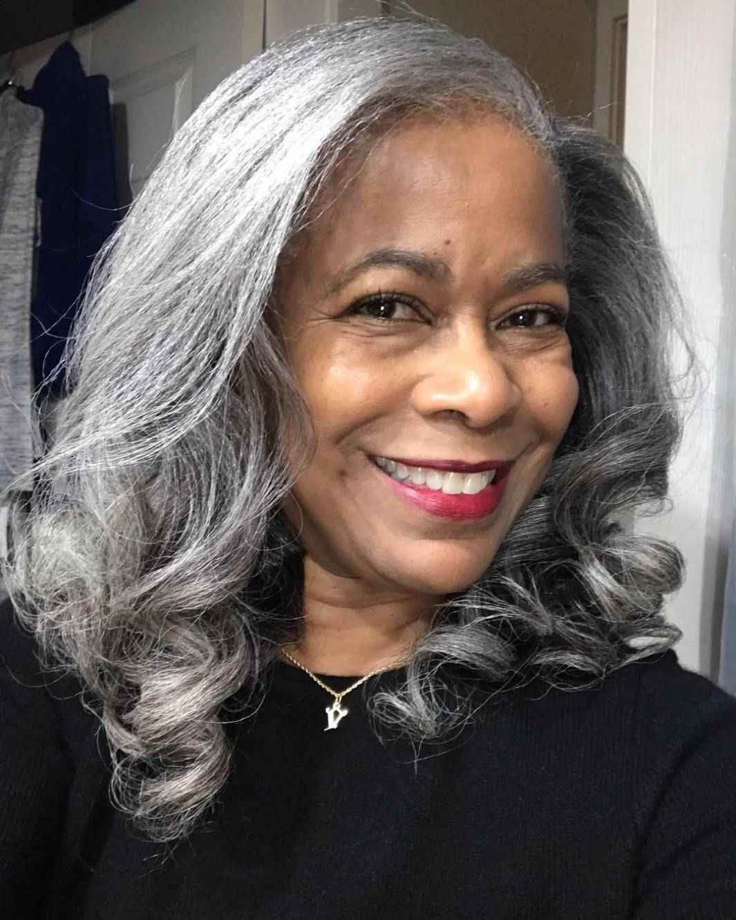 a closeup portrait of a smiling black woman with gray hair wearing a black shirt and a necklace. she looks happy and confident. her hair is styled in loose curls.