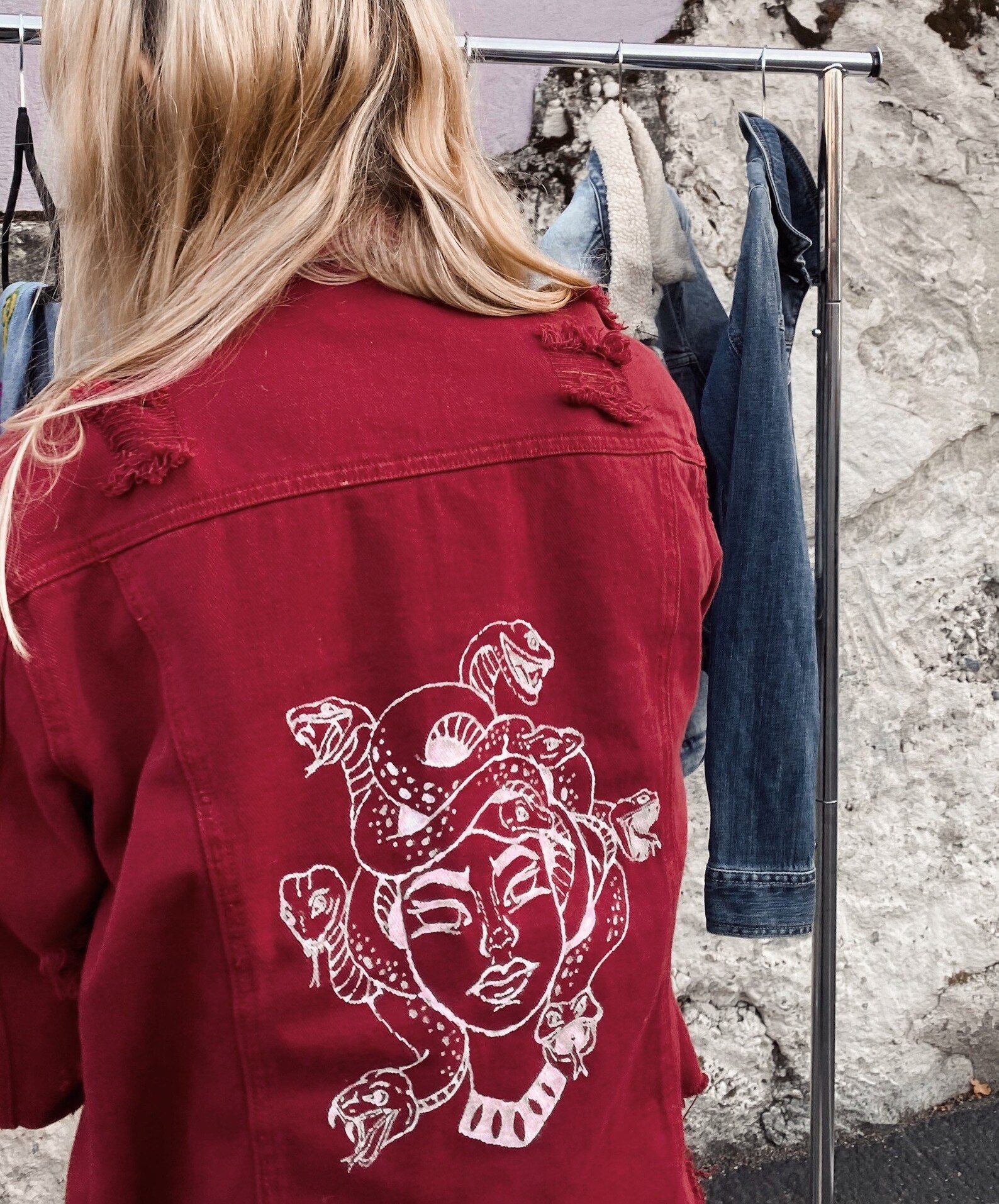 a blonde woman is wearing a red denim jacket with a white line drawing of medusa on the back. the jacket is distressed and has ripped details. the woman is standing in front of a clothing rack with other clothes hanging on it. the background is a white wall.