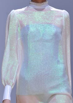 a woman wearing a white iridescent dress with long sleeves and a high neckline. the dress is sheer and has a delicate, sparkling texture. the woman's hair is out of the frame and she is standing in front of a light blue background. the dress is styled in a way that is both elegant and modern, making it a perfect choice for a special occasion.