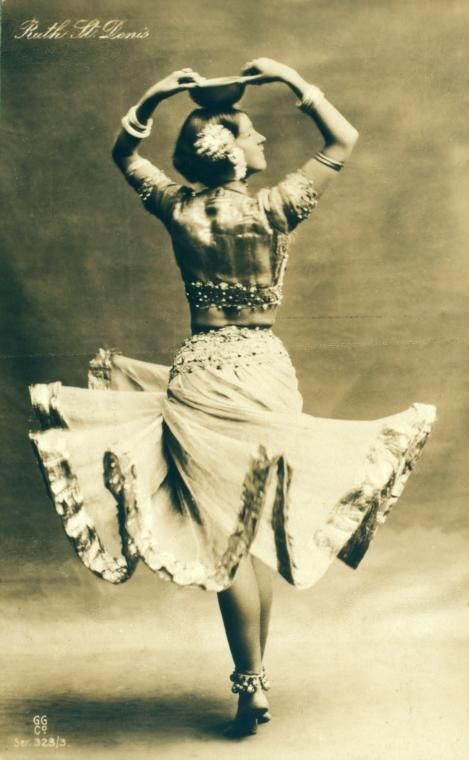 a vintage sepia photograph shows ruth st. denis performing a traditional dance. she is seen from the back, holding a bowl on her head with both hands. she has a headpiece and jewelry and wears a long, ornate dress with a bare midriff. the skirt flares out around her as she stands on one leg.
