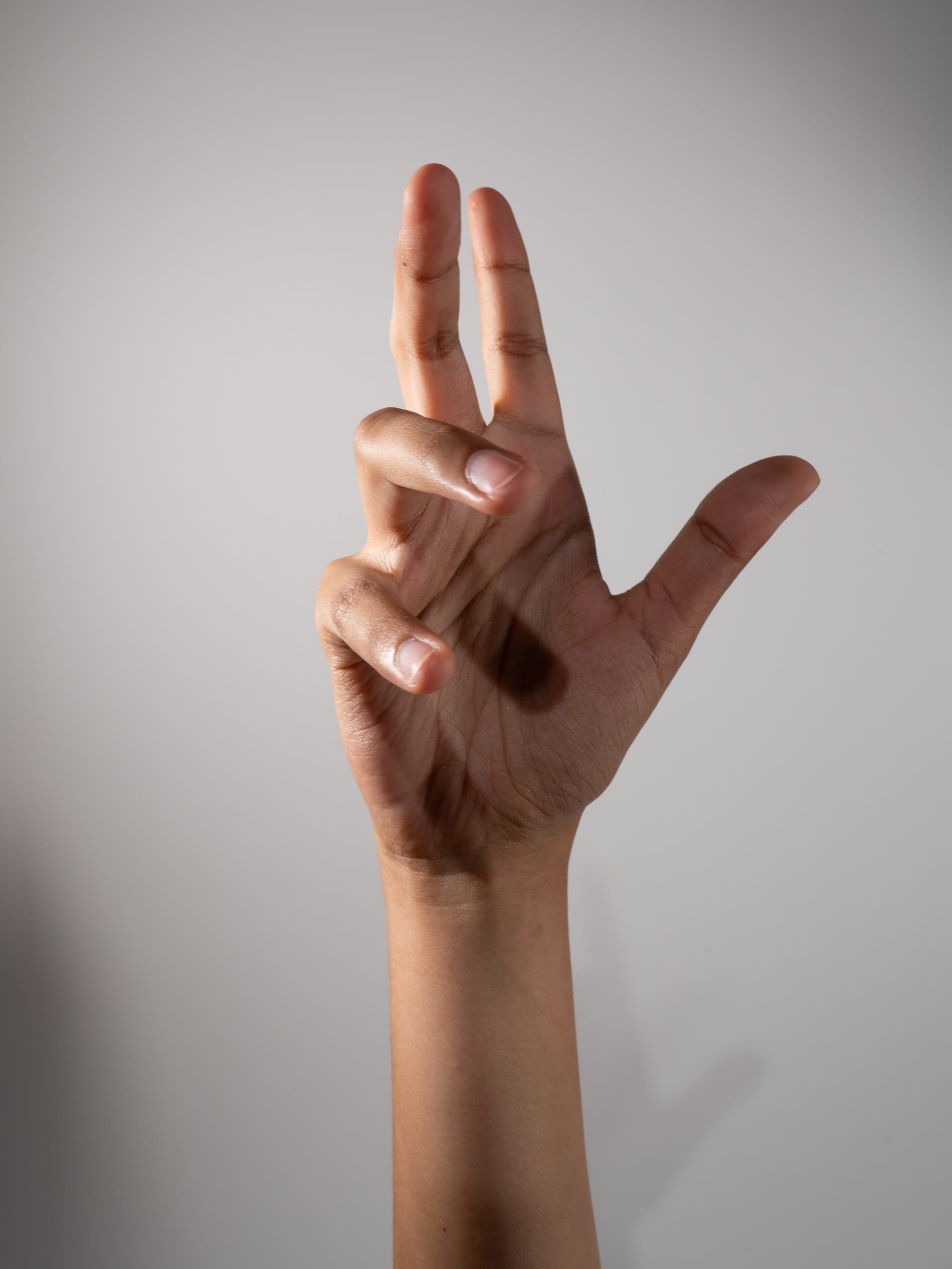 hand is extended upwards against a light grey background, displaying the number three with its fingers. the middle and ring fingers are held straight up, while the index finger is bent slightly. the pinky and thumb are folded down towards the palm. the hand is illuminated by soft light, highlighting the texture and contours of the skin.