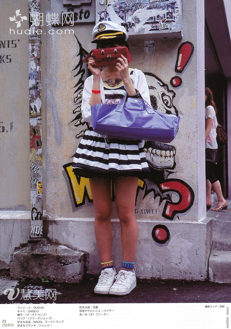 a young woman poses for a street fashion photo in japan. she's wearing a black and white striped dress, white sneakers, and a sailor hat. she's also carrying a large purple bag and holding a small red object in front of her face. the background is a grungy urban wall with graffiti and stickers.