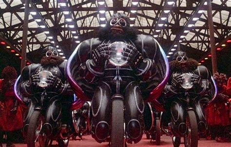 a group of bikers in futuristic leather costumes and helmets are riding motorcycles in a warehouse.  the bikers are all wearing black leather suits and helmets.  the motorcycles are all black and have a futuristic design. the bikers are riding in a formation, with the leader in the front and the others following behind.  the warehouse is dark and lit by a few lights.