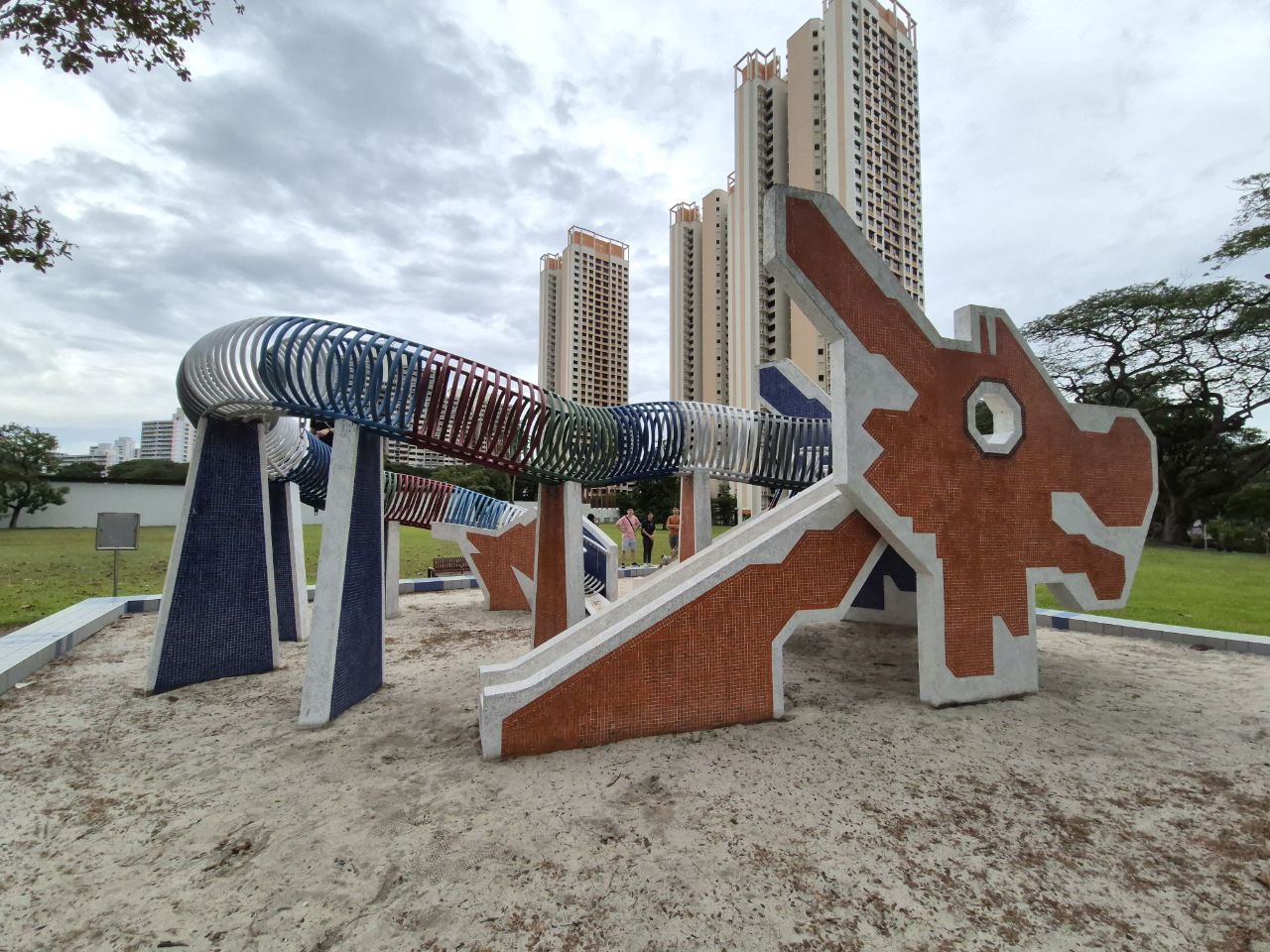 a colorful dragonshaped playground structure with a slide and a series of climbing hoops. the playground is located in a park in front of highrise apartment buildings. the playground is made of concrete and is decorated with red, blue, green, and white tiles. the dragon's head is facing the camera, and the slide is located on the right side of the dragon. the playground is surrounded by trees and grass. there are children playing on the playground in the distance. the image is taken on a sunny day with blue skies and white clouds.