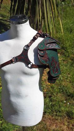 a closeup view of a beautifully crafted leather spaulder, displayed on a white mannequin. the spaulder, designed for the left shoulder, showcases intricate floral tooling and a striking combination of brown and green leather. it's set against a backdrop of lush greenery, suggesting an outdoor setting.