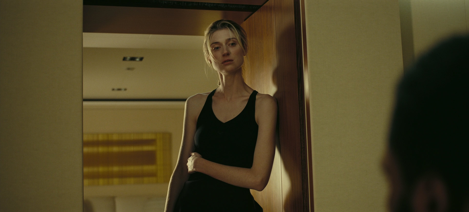 a woman with long blonde hair and a black tank top stands in a doorway, looking serious and concerned. the doorway is open, and we can see a room behind her, with light coming in from the window. she is in the shadows, but her face is illuminated. the woman's expression suggests that she is worried or sad. she is standing in a doorway, which could symbolize a transition between two states of being. the lighting and shadows add to the dramatic mood of the image. this looks like a film still from a movie.