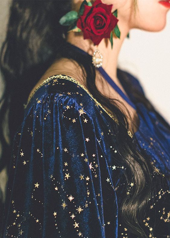 the image shows a closeup of a woman wearing a blue velvet dress with gold stars. the dress has a halter neckline and puffy sleeves. the woman has long black hair and is wearing a red rose behind her ear. the lighting is soft and the perspective is a closeup, focusing on the details of the dress and the woman's profile.