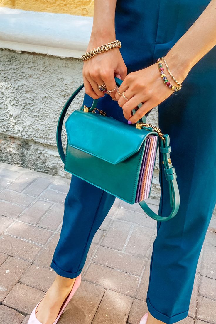 a closeup shot of a woman wearing a teal pantsuit and carrying a teal handbag with a multicolored stripe detail on the side. she is wearing gold and colorful bracelets and rings. the photo is taken outdoors on a pavement.