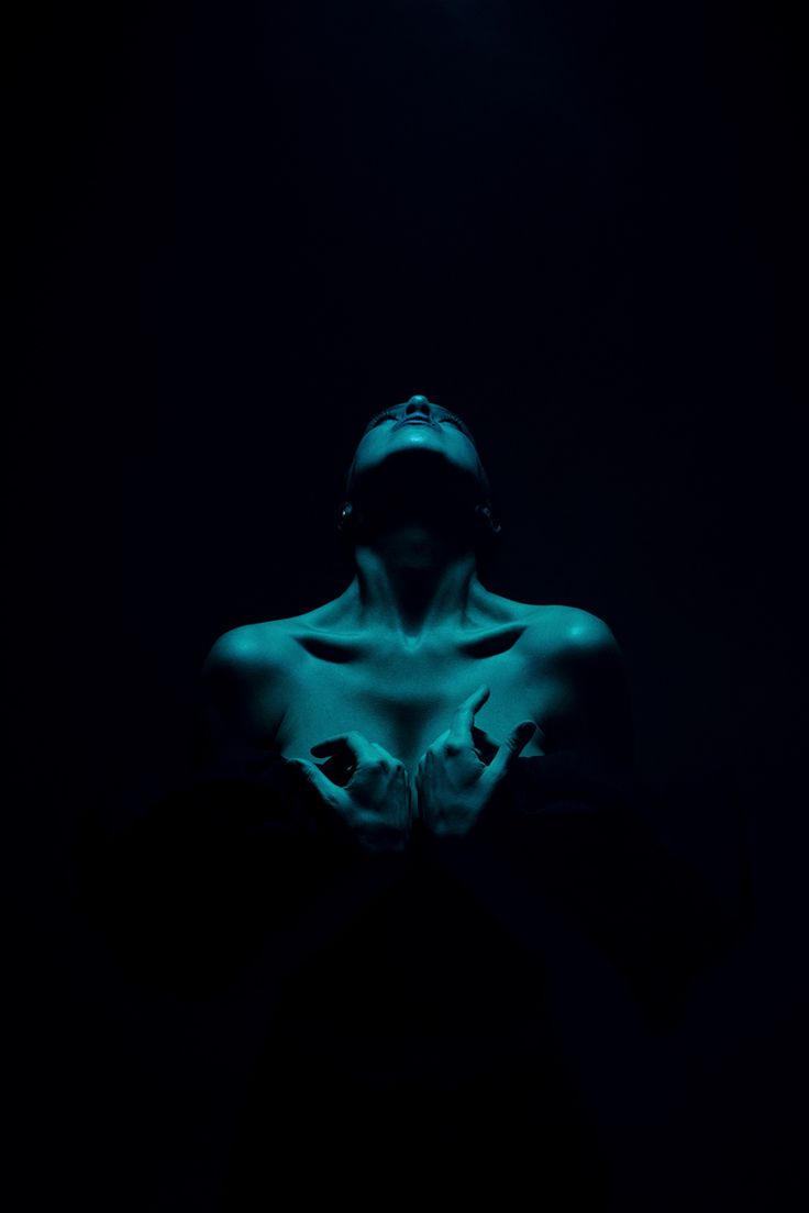the image is a stylized portrait of a barechested woman. her body is mostly in shadow, with only her torso and head visible. the woman is positioned facing upwards, her hands clasped near her chest. a strong, cool blue light illuminates her, creating a dramatic contrast with the deep black background. the overall mood is dark, sensual, and mysterious. the photo's composition and lighting create a feeling of intimacy and vulnerability.