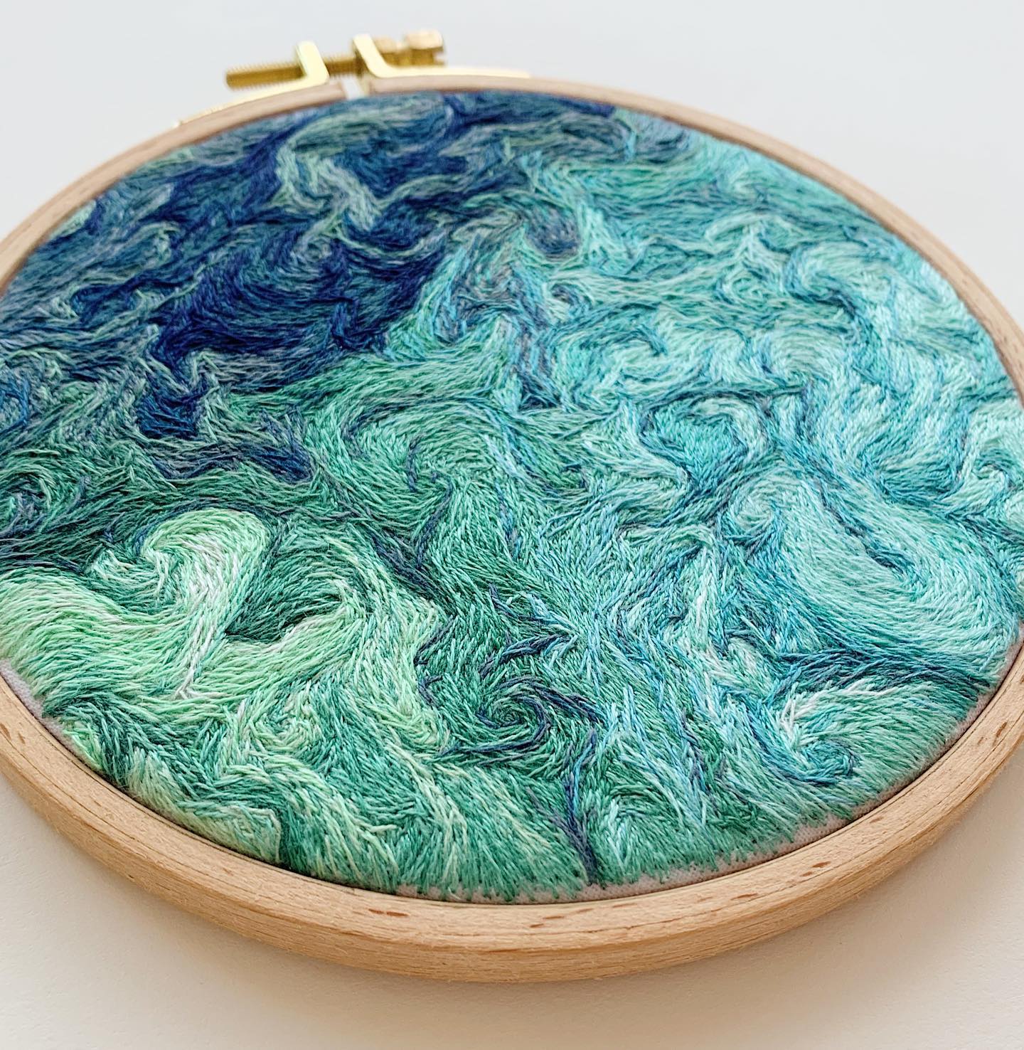 the image showcases a piece of modern embroidery art. an embroidery hoop holds a fabric adorned with an abstract design of ocean waves. the artwork utilizes a captivating blend of blue, turquoise, and green hues, skillfully crafted through thread work to capture the fluidity and depth of the sea. the closeup perspective highlights the intricate details and texture created by the embroidery stitches.