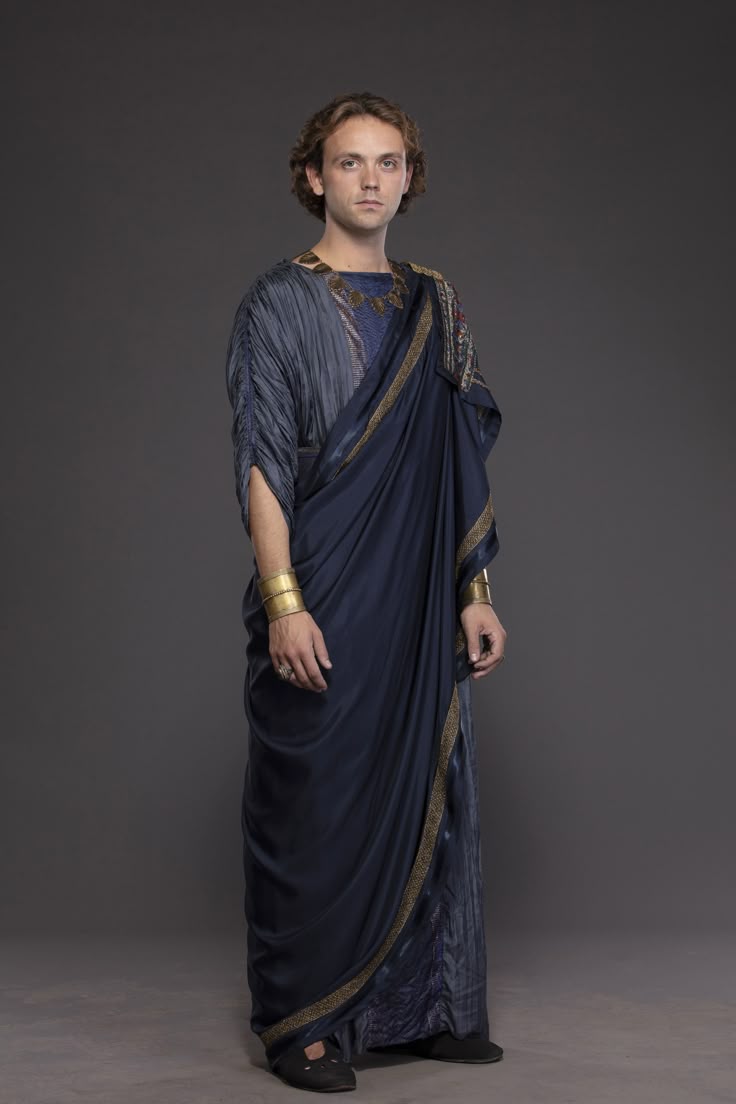 a fullbody portrait shows a man in an ancientstyle tunic with gold bracelets on his wrists and rings on his fingers. he has curly brown hair, and he looks directly at the viewer. the tunic has different colors and materials, with the top part being grayblue and the lower part being dark blue with gold trim. his jewelry is visible at the neckline. he is standing against a gray background, and black shoes are visible at the bottom of the frame.