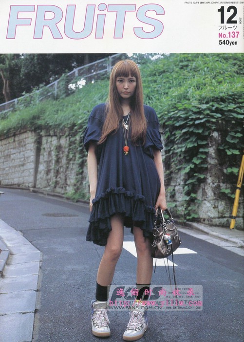 a young woman with long brown hair and bangs poses on a city street for the cover of fruits magazine. she's wearing a navy blue ruffled dress, white hightop sneakers, and black socks. she carries a silver handbag with a long strap. the background features a stone wall with greenery and a sidewalk. the lighting is natural daylight, giving the image a slightly overcast look.