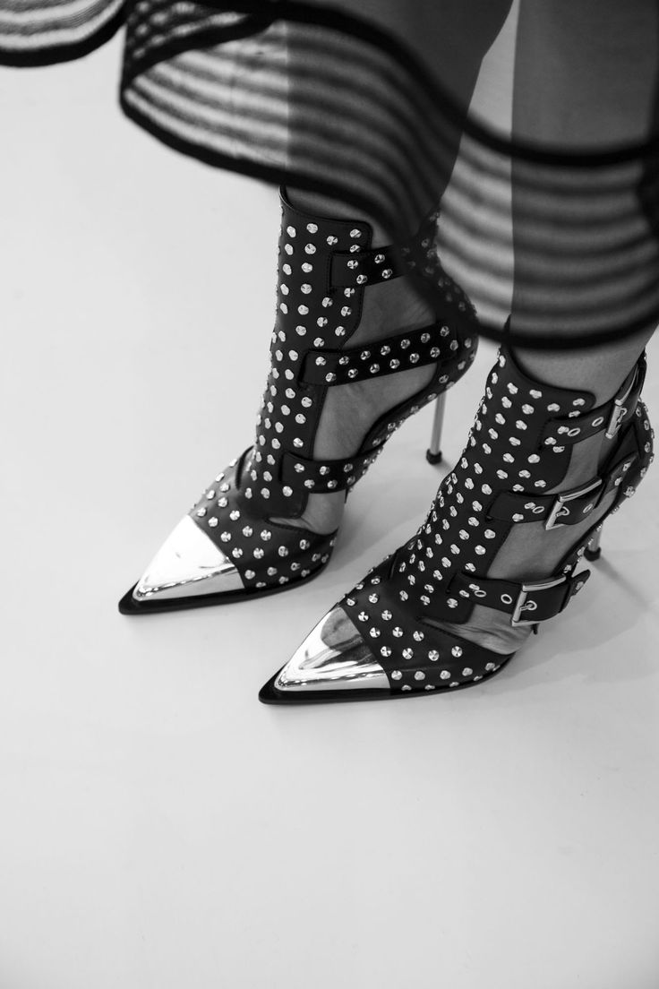 a black and white image of a woman wearing a pair of high heeled black leather boots with silver studded embellishments. the boots are opentoed and have a pointed toe. the image is taken from the side and focuses on the boots, with only a small part of the woman's legs visible. the background is a simple white surface.