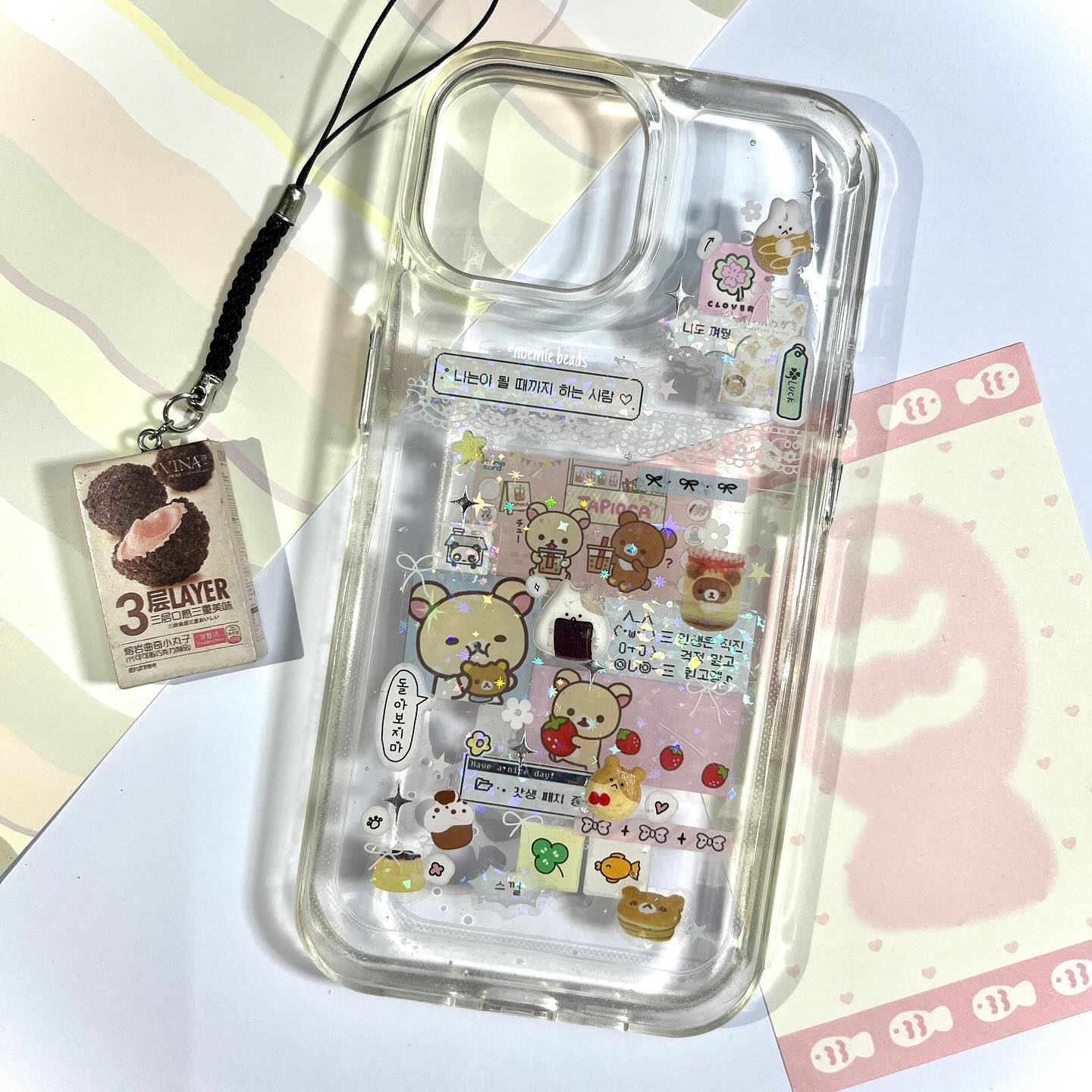 the image features a clear phone case decorated with various stickers and a charm. the phone case has a transparent lanyard with a small boxshaped charm attached. the charm is labeled "3 layer" and displays an image of chocolate balls. inside the phone case, there are an assortment of cute stickers, including cartoon bears, food items, and handwritten phrases in korean. to the right of the phone case is a pink and white card with a pattern of fish. the background includes white and lightcolored surfaces, providing a clean and bright setting for the phone case and accompanying items.
