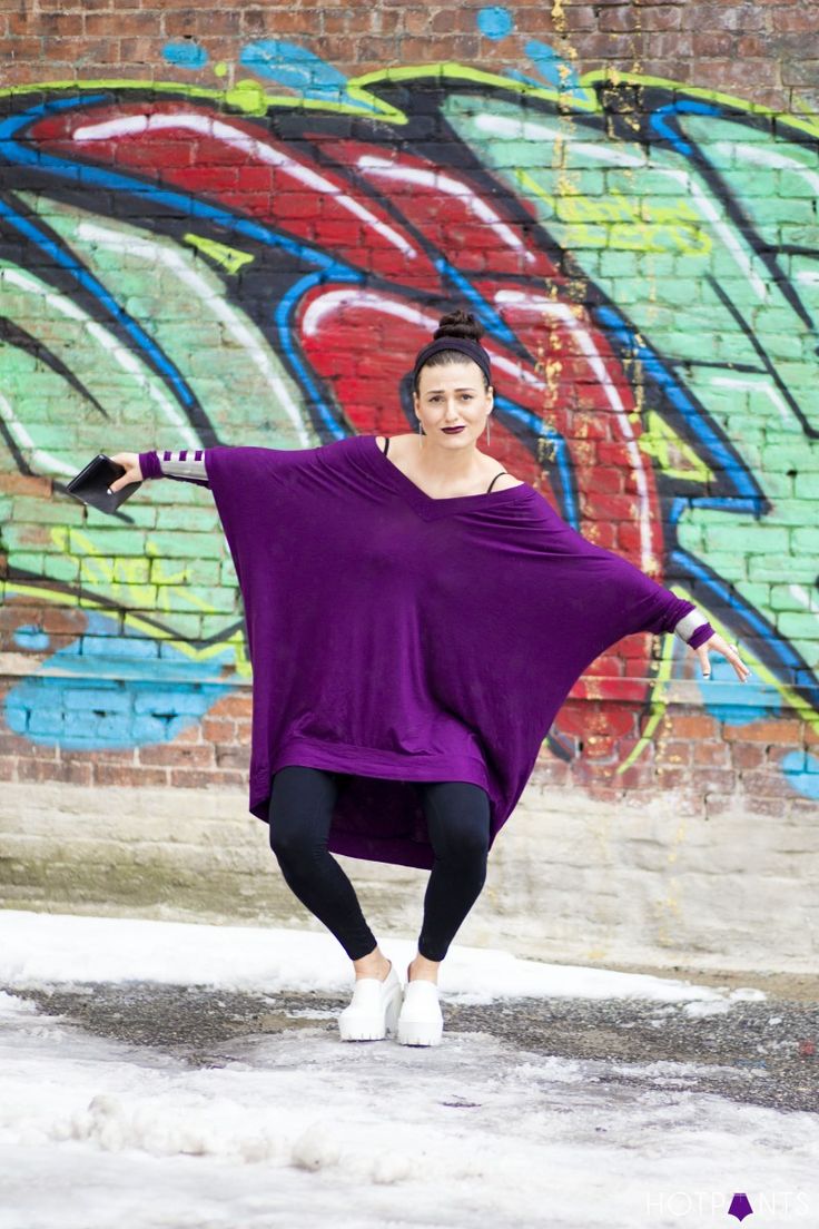 a woman in a purple tunic, leggings, and white platform shoes strikes a pose against a graffiticovered brick wall. she is holding a tablet in one hand and has her arms outstretched. the setting appears to be outdoors with some snow on the ground.