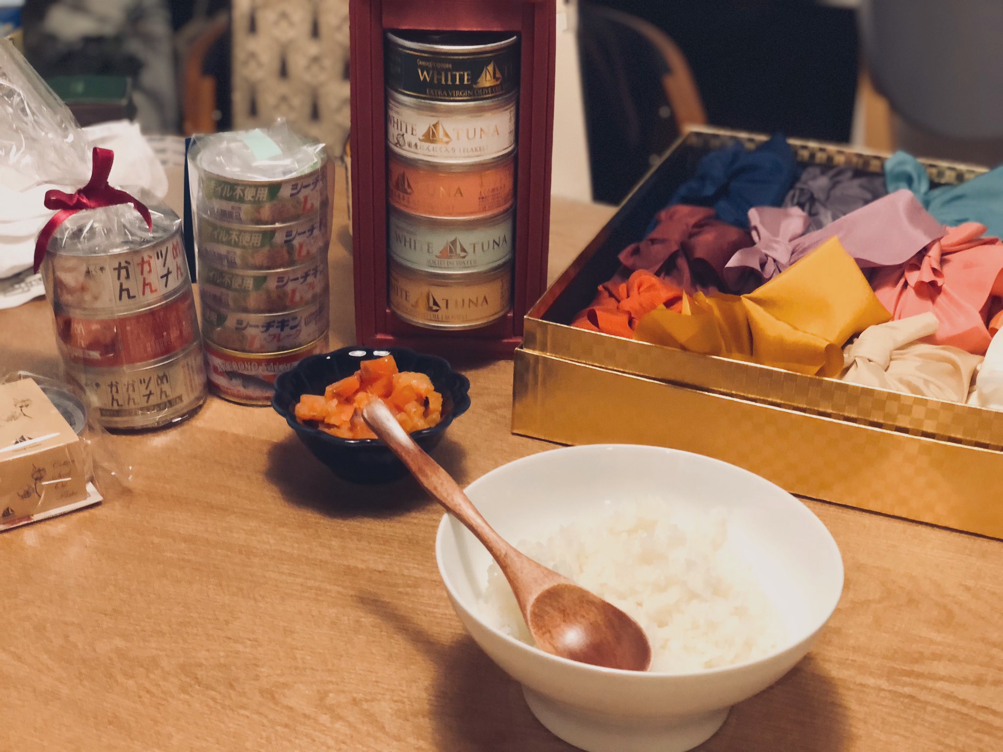 a wooden table is laid out with various snacks, including cans of tuna stacked in a pyramid shape, a bowl of pickled ginger, and a bowl of white rice with a wooden spoon. there's also a variety of colorful fabrics on the table.