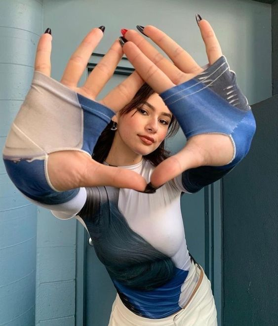 a woman is taking a selfie, making a triangular shape with her hands in front of her face. she is wearing a white and blue longsleeved shirt with an abstract print and white pants. her nails are painted black. the background is a light blue wall and the photo appears to be taken indoors under artificial lighting.
