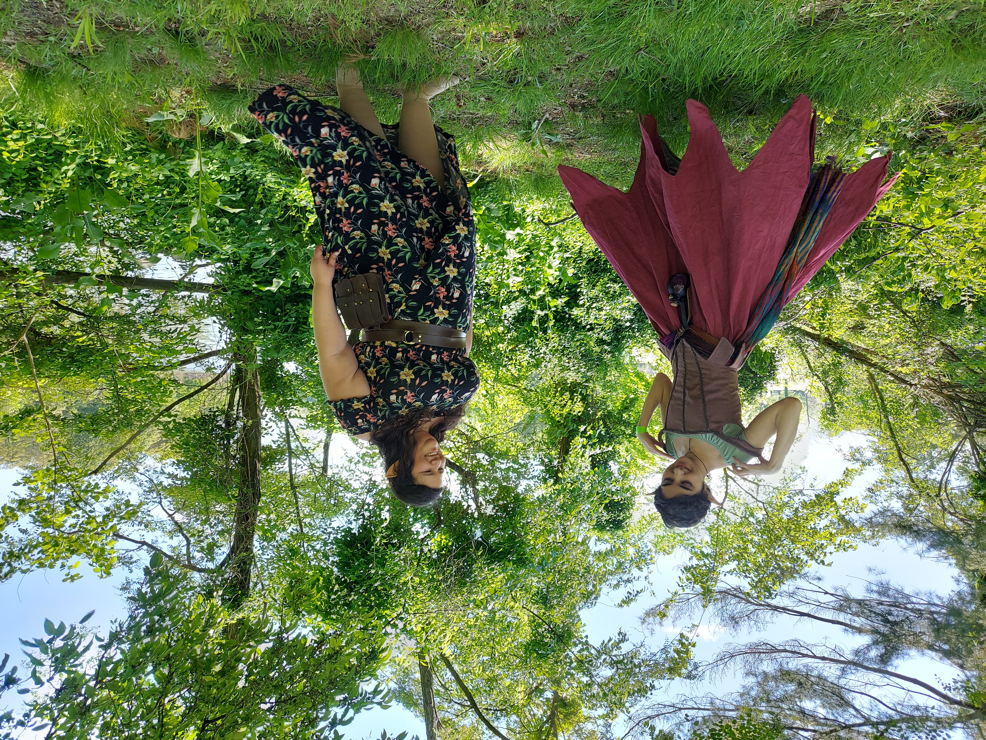 two women, one dressed in a whimsical, pointedhemmed maroon dress and corset resembling an elf costume, and the other in a floral maxi dress, stand in a lush green forest near a river.  the woman in the elf costume is adjusting her dress, and they both appear to be enjoying the natural setting. the overall mood is whimsical and fantastical.