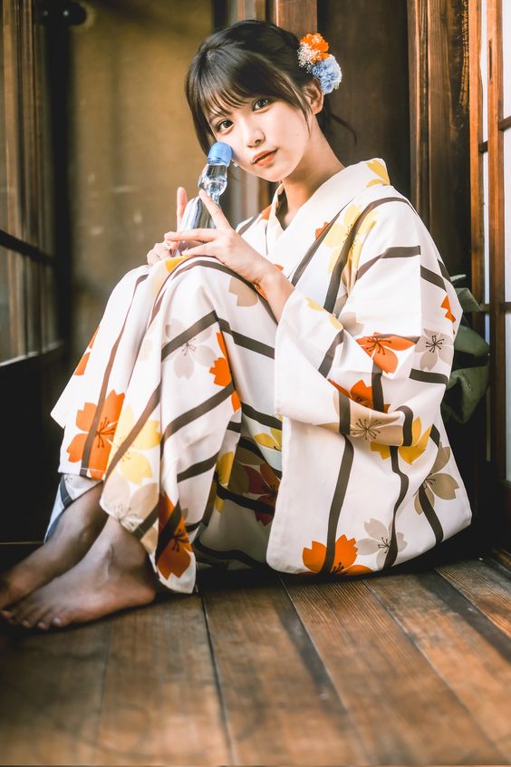 a young woman in a colorful yukata (summer kimono) sits gracefully on a wooden floor. she's holding a glass bottle, possibly containing a refreshing drink, and gazes directly at the viewer. her hair is styled with a blue flower ornament, adding a touch of elegance. the setting appears to be a traditional japanese house, evident from the wooden architecture and shoji screen in the background. the soft, natural lighting creates a warm and inviting atmosphere.
