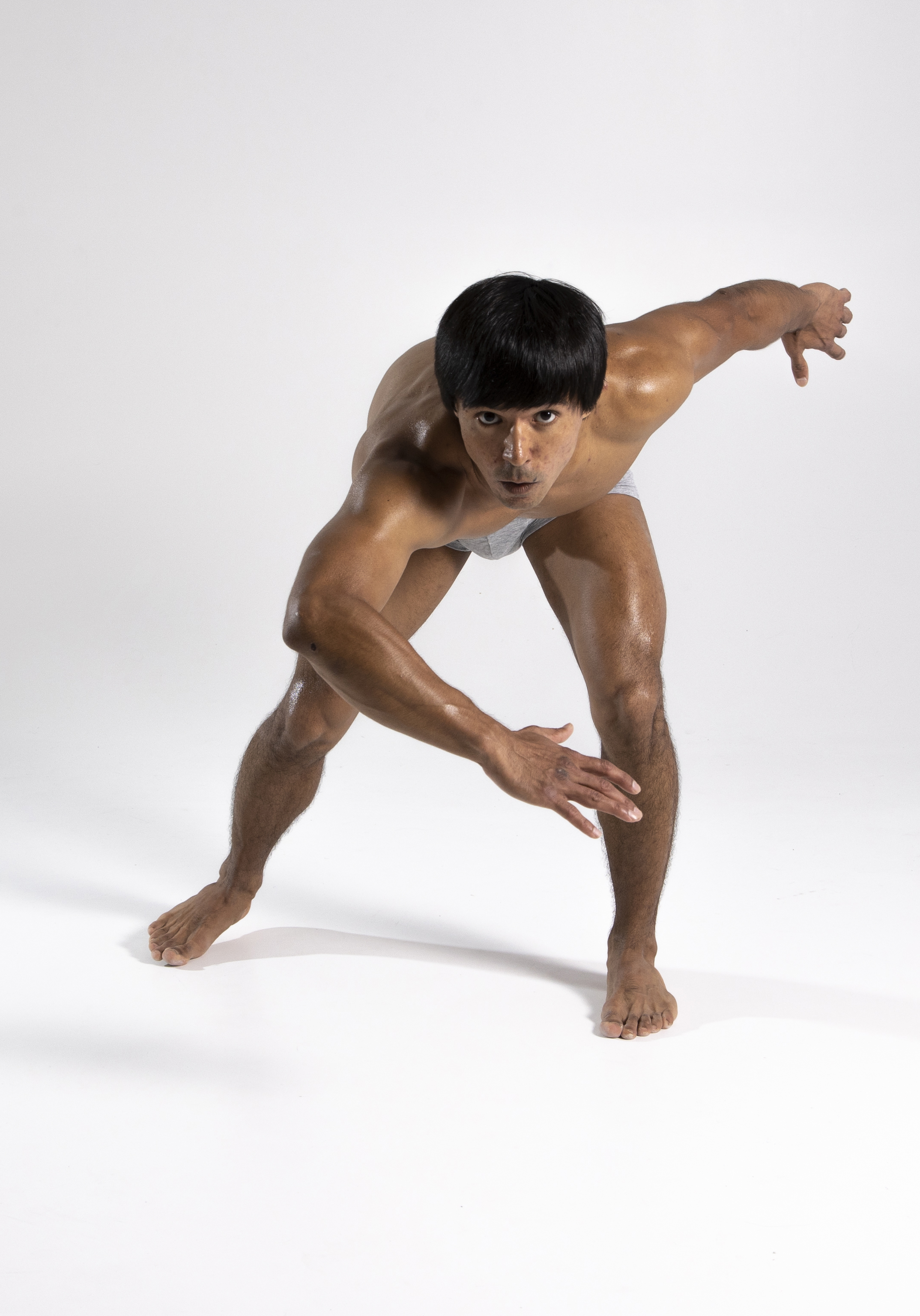 a muscular, athletic asian man with dark hair is captured in a dynamic pose in a studio setting. he is mostly bare, wearing only underwear, and is barefoot. the man is leaning forward with one arm extended out and the other arm bent and positioned across his body. his intense gaze is directed at the camera. the bright, white background emphasizes the man's physique and the energy of his pose.
