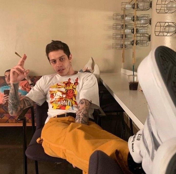 a young man with tattoos on his arms and legs, is sitting on a black chair, wearing a white tshirt with a colorful graphic design, yellow pants, and white sneakers. he is holding a cigarette in his right hand, looking at the camera with a smile.  a black cup of coffee is on the counter behind him. he is casually dressed and appears relaxed.