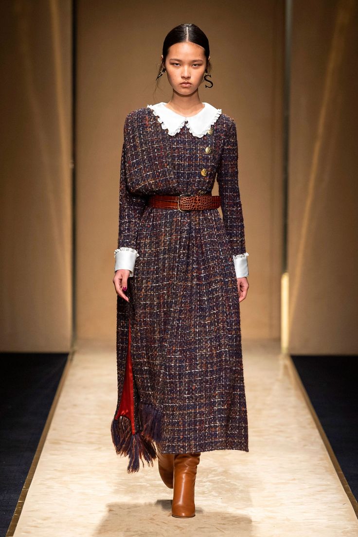 a full shot of a fashion model walking down a runway. she is wearing a longsleeved midi tweed dress with a peter pan collar and a brown leather belt. the dress is a dark navy and burgundy color scheme with a subtle checkered pattern.  she's also wearing brown leather boots and carrying a small, fringed handbag with a red interior visible. the backdrop is a beige runway with muted background lighting. the overall style is elegant and sophisticated.