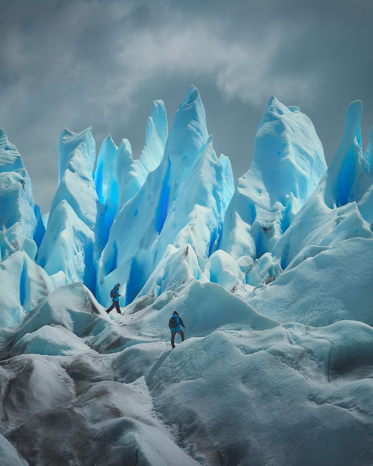 the image shows two people hiking on a large glacier. the glacier is made up of different shades of blue and grey ice formations. in the background are tall, jagged ice peaks and a cloudy grey sky.