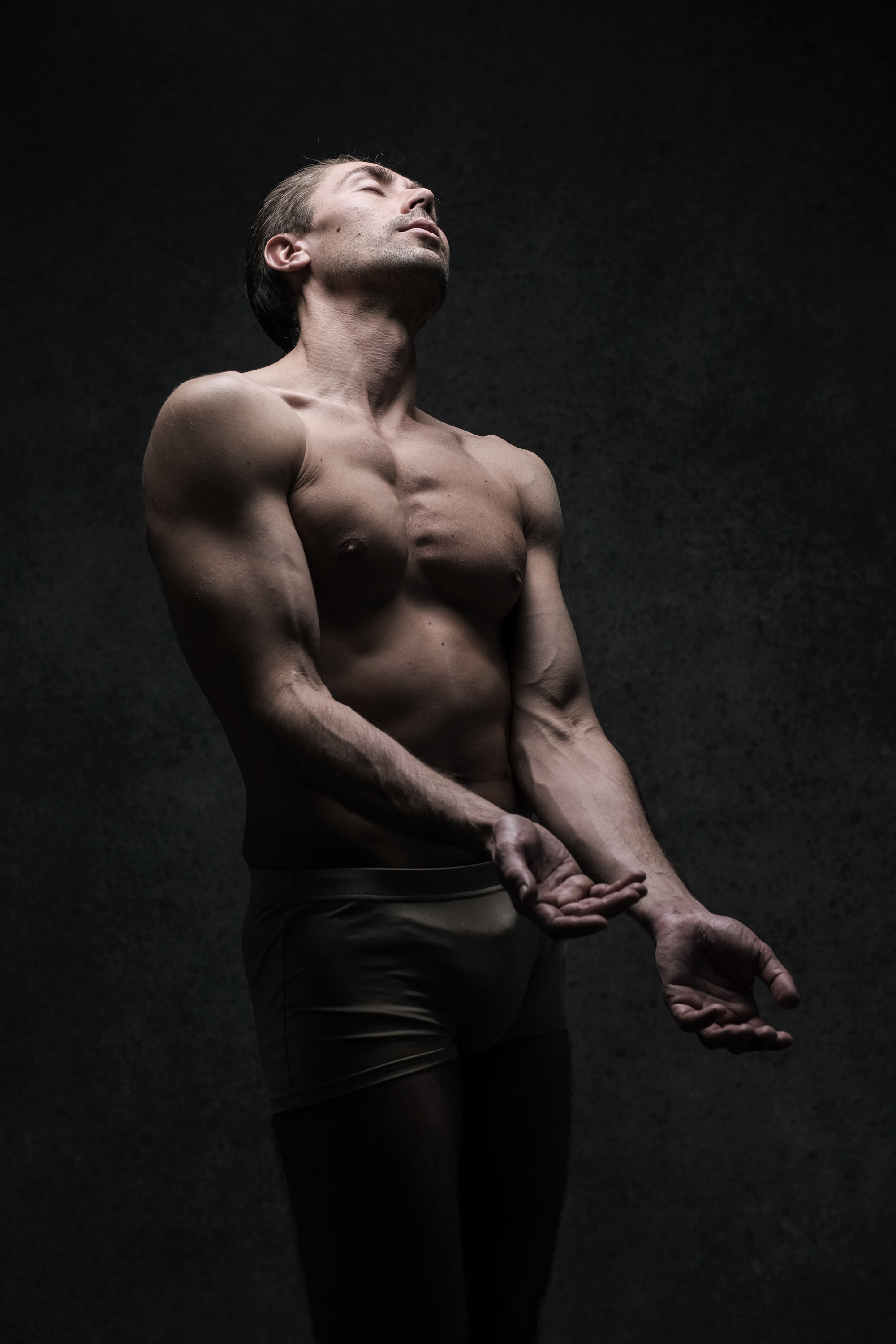 a muscular man stands with his head tilted back and eyes closed, arms extended in front of him, palms open, as if in a state of surrender or ecstasy. his physique is illuminated by a spotlight against a dark, textured background, creating a dramatic and evocative image.